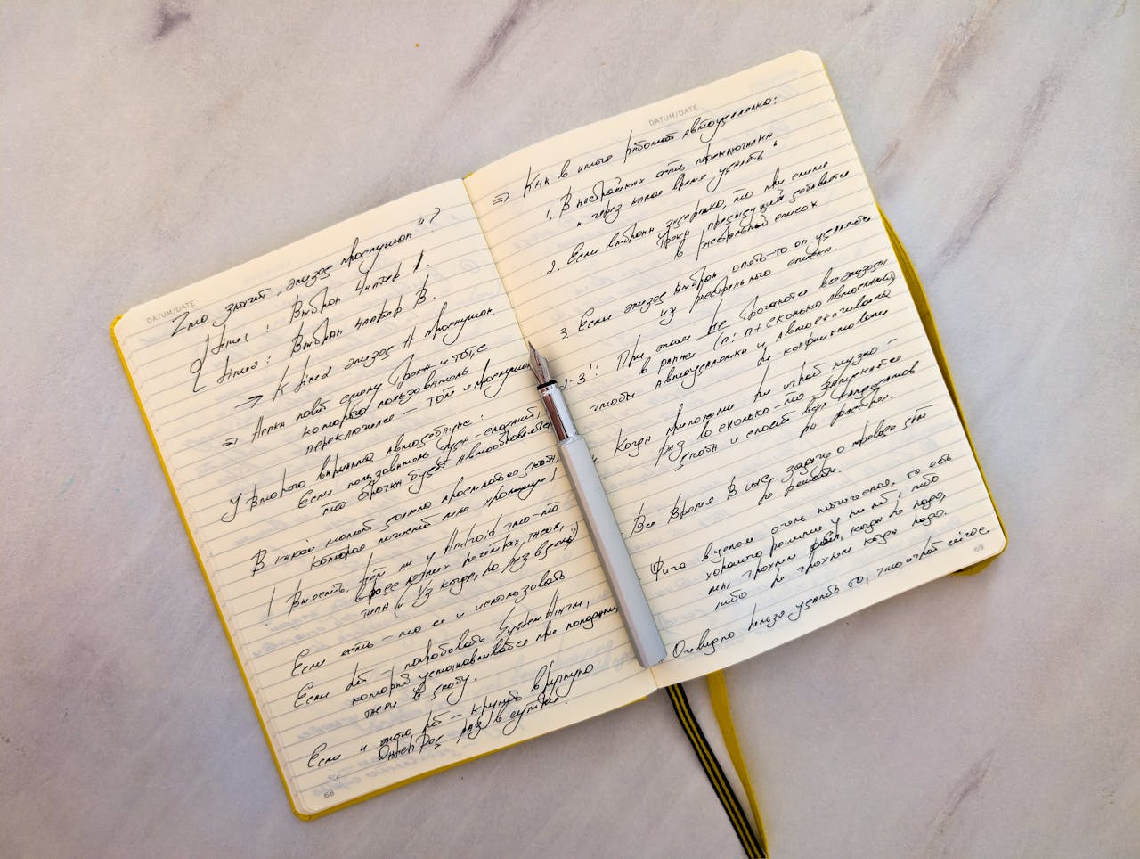 Open notebook with cursive handwritten notes on marble surface and a pen resting on pages.